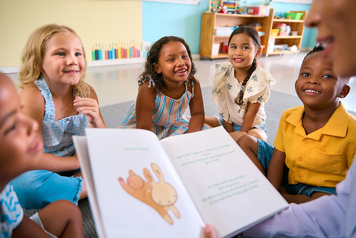 Female Primary Or Elementary School Teacher Reads Story To Multi-Cultural Class Seated In