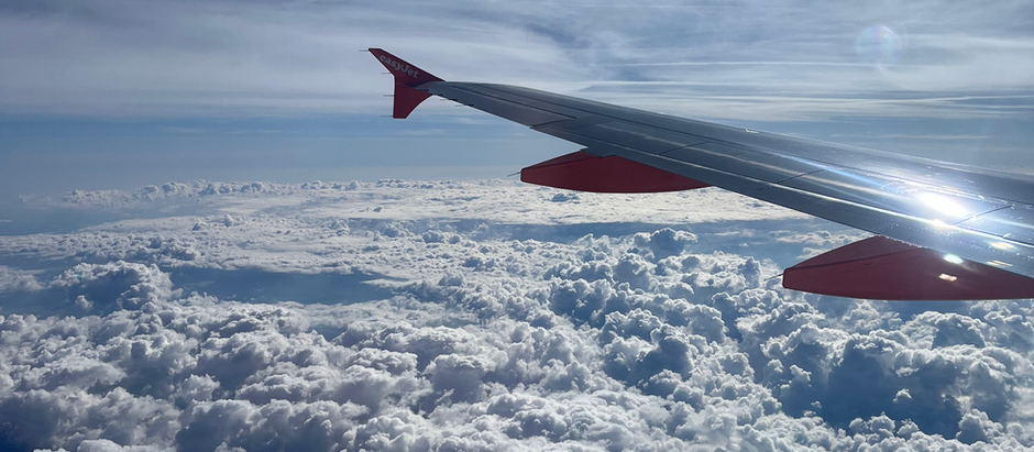 Airplane wing with red tip gliding over fluffy clouds under a bright blue sky. Sun beams and contrails are visible, evoking tranquility.
