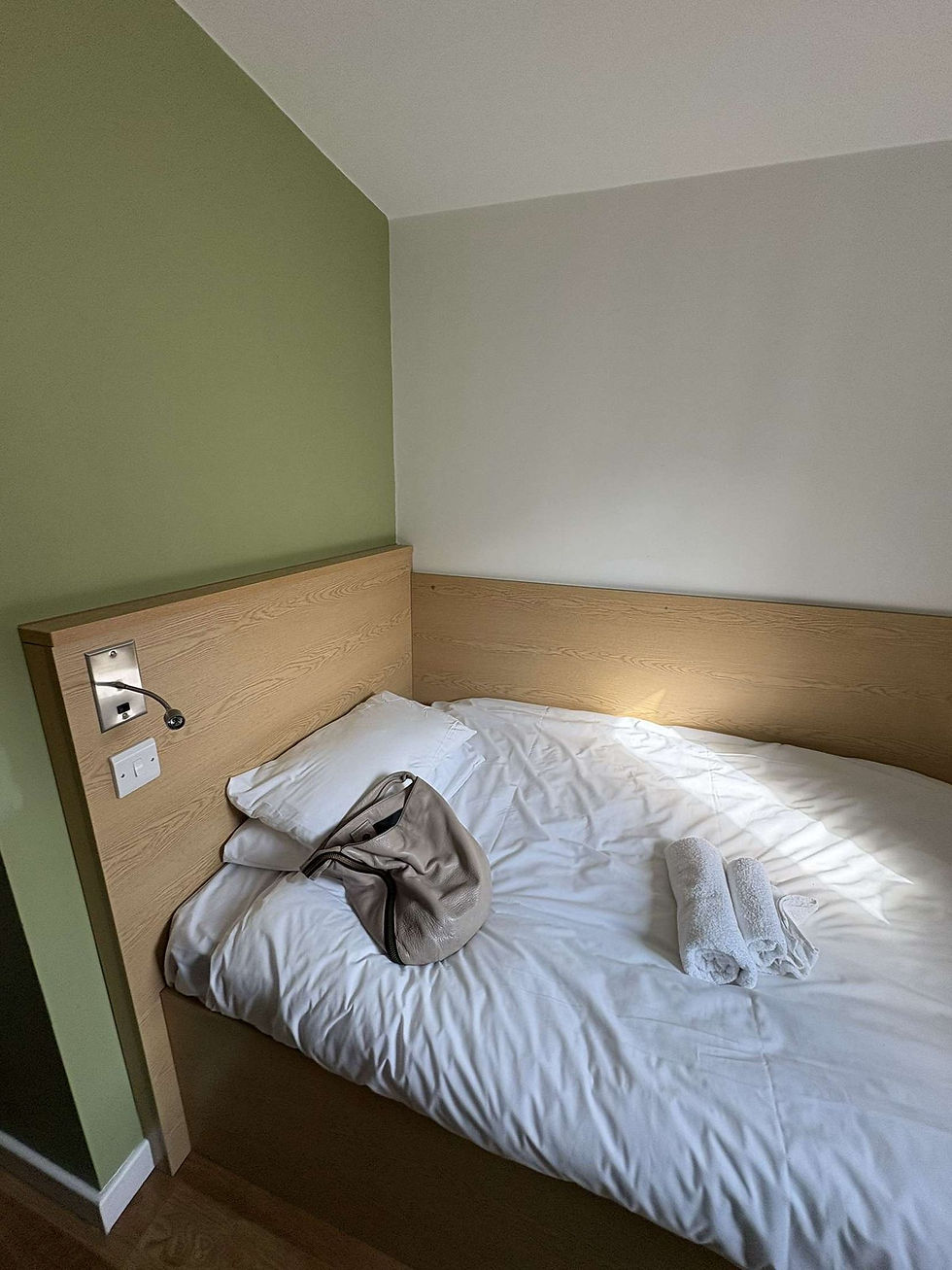 A neatly made bed with white sheets, a beige bag, and rolled towels in a room with green and white walls, creating a tidy and calm atmosphere.