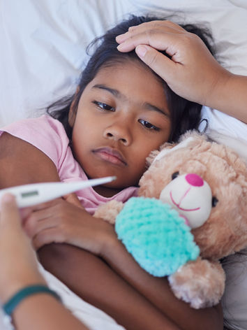 A child in bed holding a teddy bear, her mom's hand on her forehead. Her mom's other hand is visible holding a thermometer
