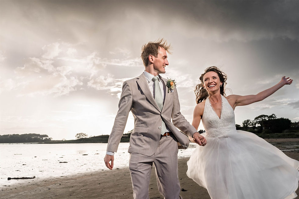 Bride and groom joyfully run on a beach. Bride in a white dress, groom in a grey suit. Dramatic cloudy sky in the background.