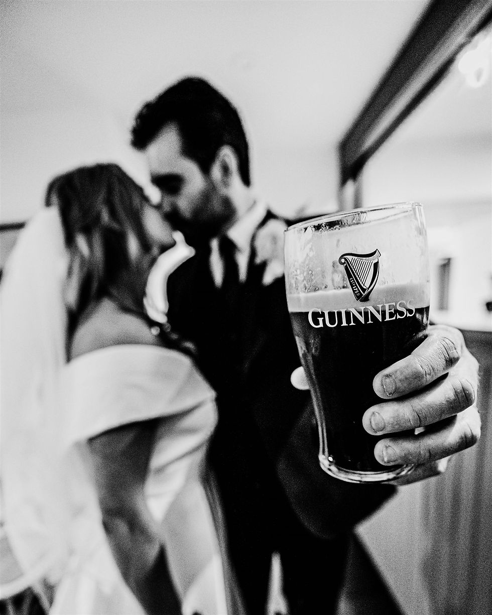 Killeavy Castle Wedding Photography - Bride and groom kissing, with a close-up of a hand holding a Guinness pint glass in the foreground. Black and white, wedding setting.