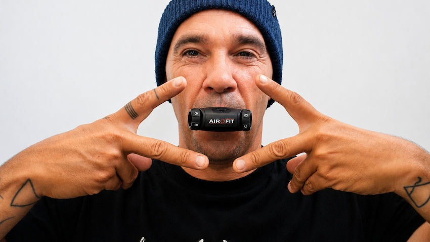 Close-up of a freediving instructor in a navy beanie holding fingers near his face around an AIROFIT mouthpiece, against a white background.