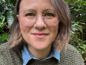 Smiling woman with short hair and round glasses wearing a green sweater and denim shirt.
