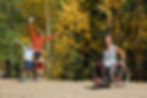 In a yellow-green forest, Ryan sits in her wheelchair and smiles at the camera while her smiling husband and two children wave with outstretched arms.