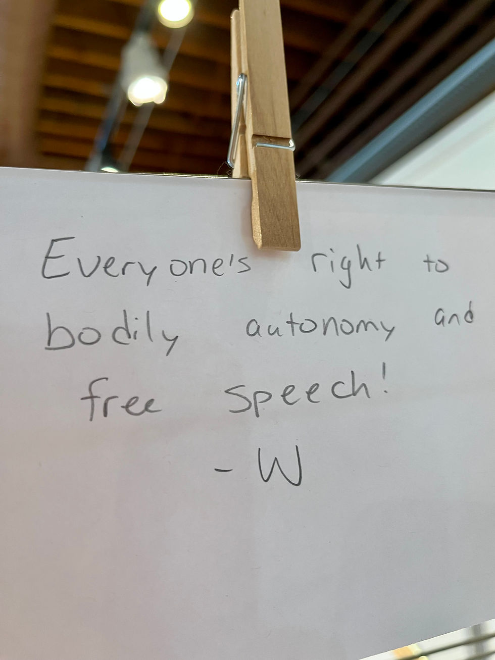 Paper with handwritten message on bodily autonomy and free speech clipped to a string. Ceiling lights visible in background.