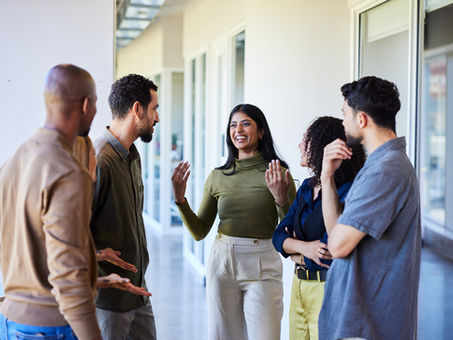 A diverse group of colleagues standing in a hallway, engaged in a lively conversation, with one woman speaking and others listening attentively.