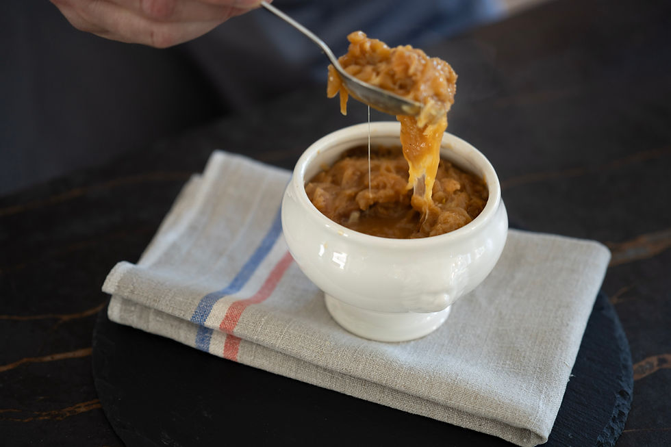 A hand holds a spoon over a white bowl of French onion soup, with melted cheese, on a folded napkin with red and blue stripes.