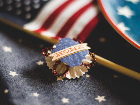 Small HOME badge on star-patterned fabric beside a patriotic American flag plate, warm festive red, white and blue mood