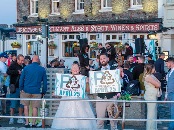 People holding "Awareness Day April 25" signs outside a pub with a busy crowd and band. The scene is festive and lively.