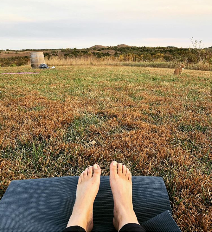 A pair of feet on a yoga mat overlooking a vineyard and hilly landscape.