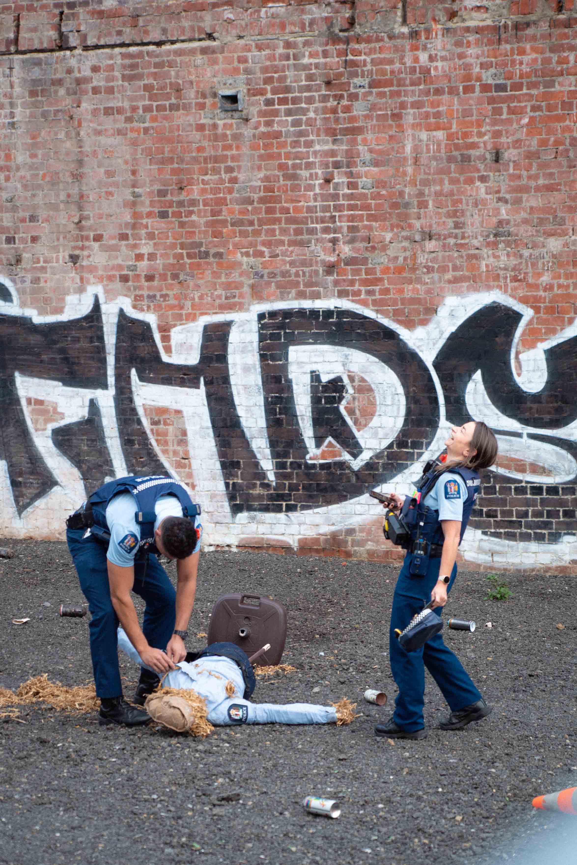 New Zealand police officers respond to Cameron Hunt’s scarecrow installation, shown laughing and inspecting the dismantled scarecrow lying on the ground. The playful interaction between real officers and the straw-filled figure highlights themes of authority, absurdity, and public space.