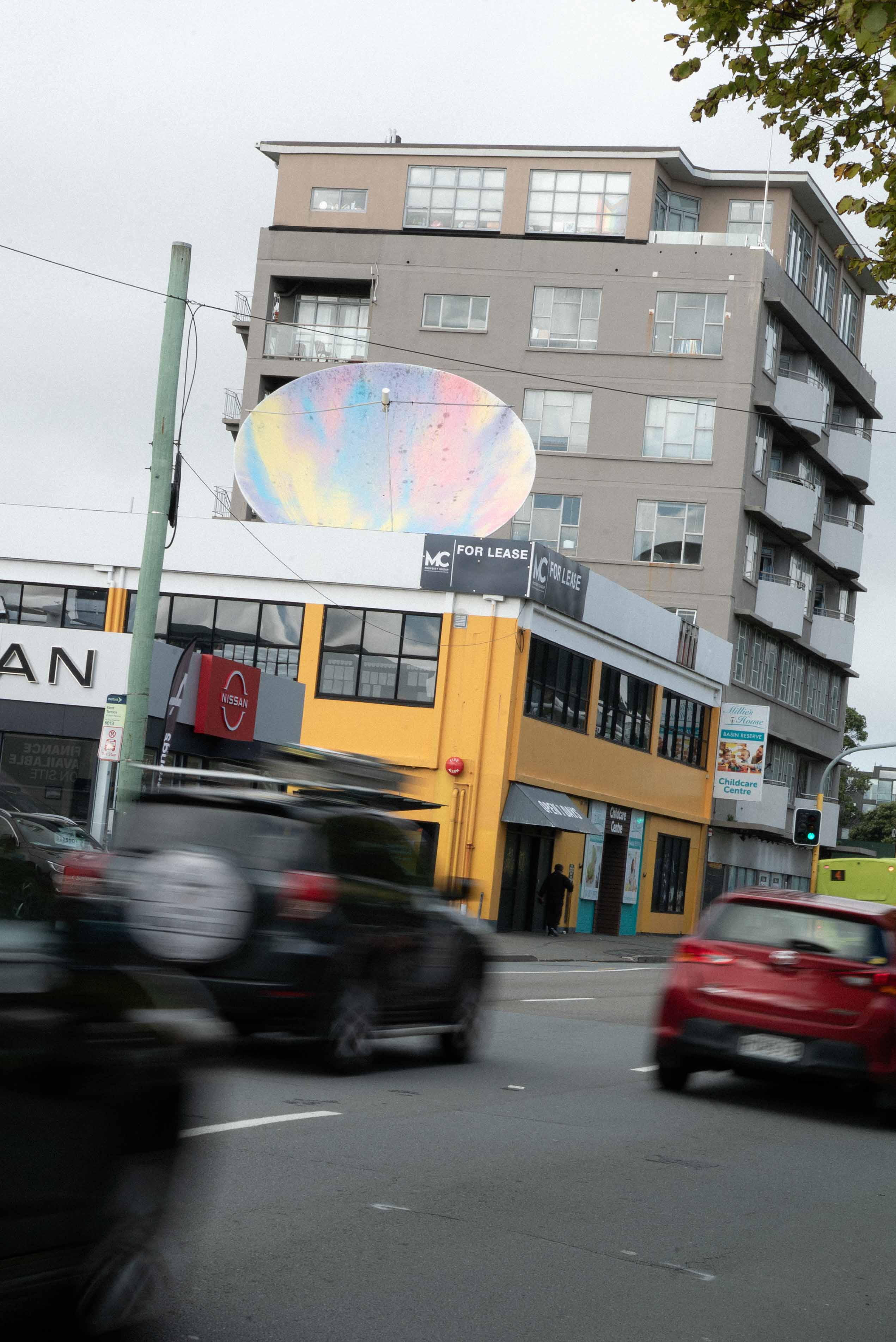 Street art installation by Cameron Hunt featuring a large rooftop graffiti piece painted on a satellite dish with soft pastel hues of blue, pink, yellow, and purple, transforming a commercial building in the cityscape.