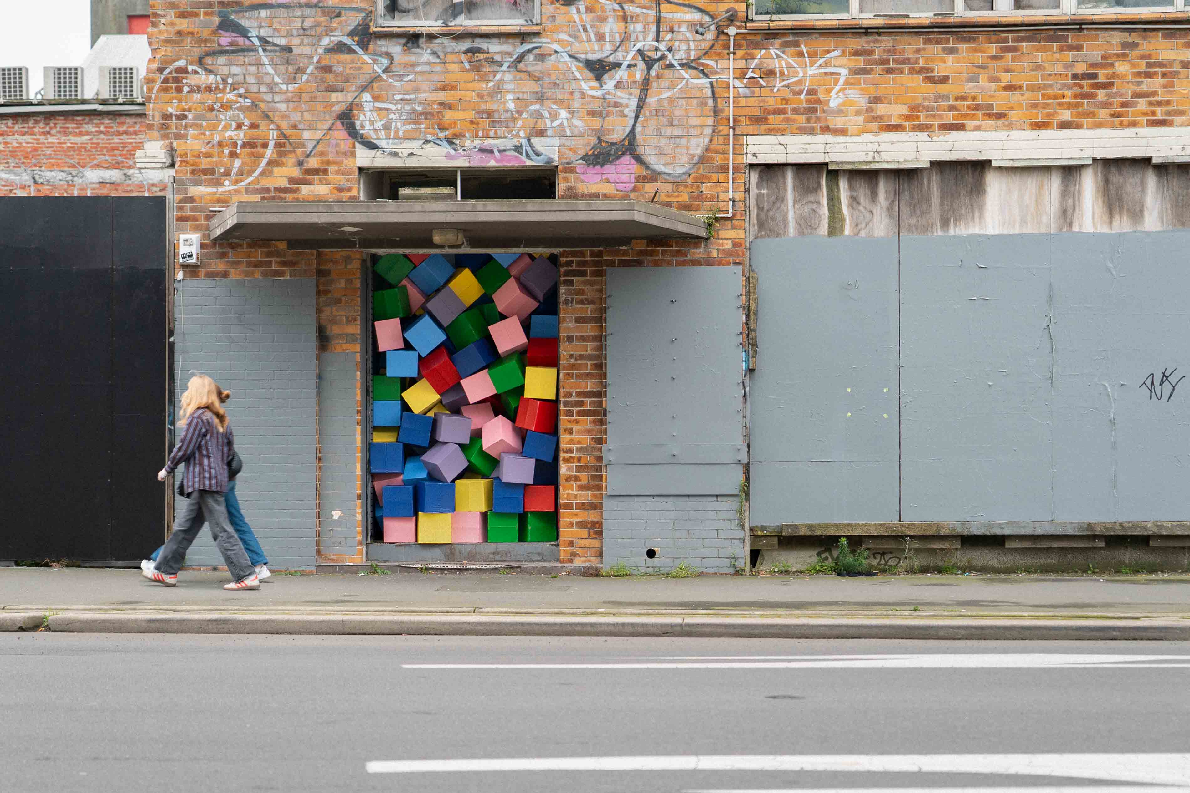 Street installation by Cameron Hunt featuring brightly coloured cardboard boxes tightly packed into a city doorway, transforming an abandoned entrance into a playful, geometric explosion of form and colour.