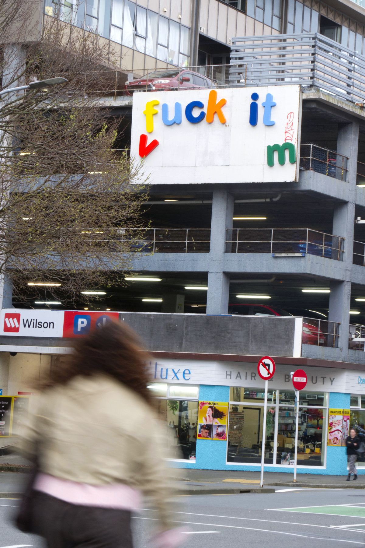 Outdoor street art installation by Cameron Hunt featuring colorful giant fridge magnet letters on an urban billboard overlooking passing cars and pedestrians on a city street.