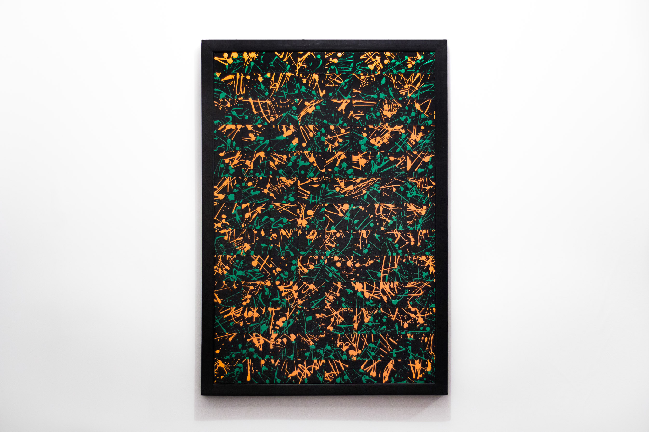 Framed abstract painting featuring layered green and orange acrylic marks over a black background. The composition includes splashes, drips, and angular linework arranged in a dense, chaotic pattern. Set in a matte black rectangular frame.