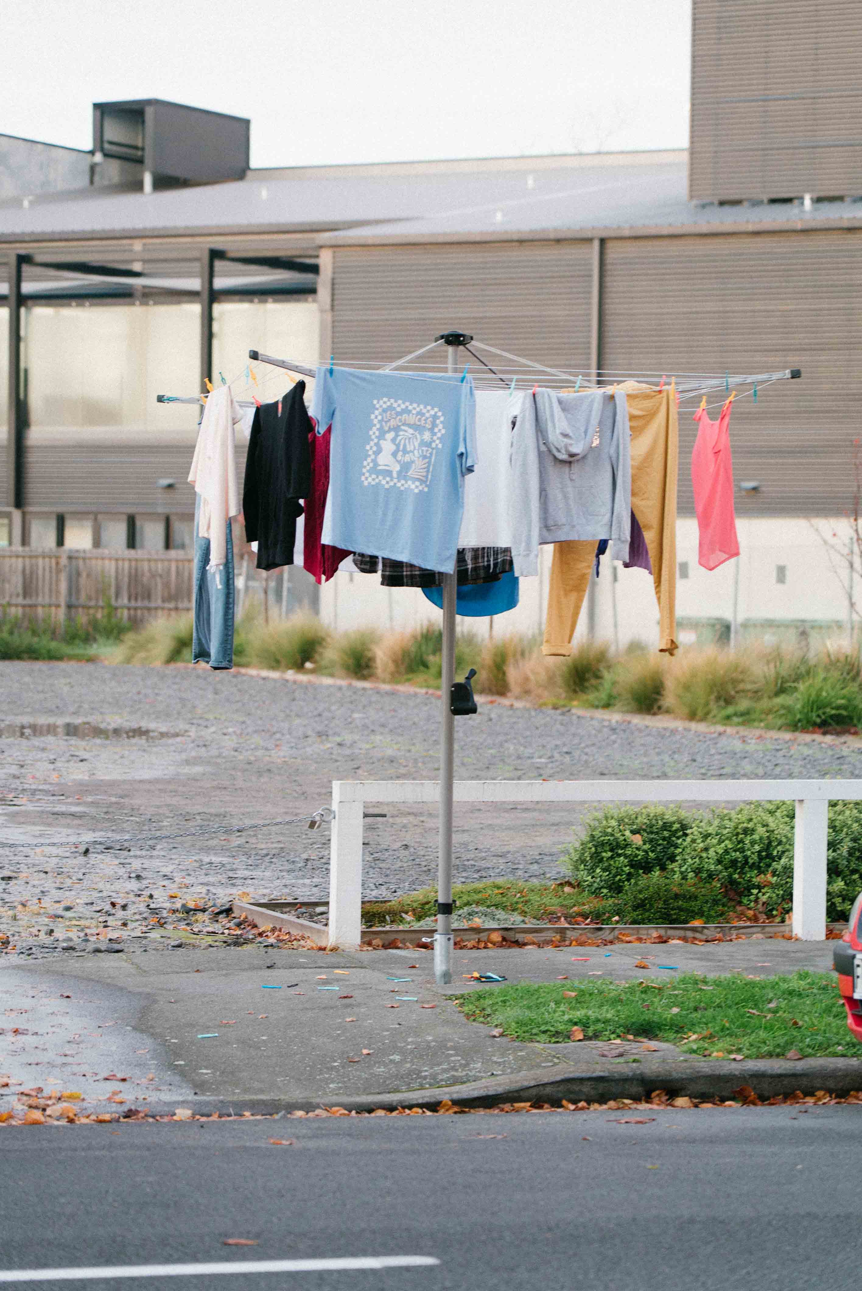 Street installation by Cameron Hunt featuring a rotary clothesline bolted into a pavement hole in the city, complete with real clothing hanging as if from a suburban backyard.