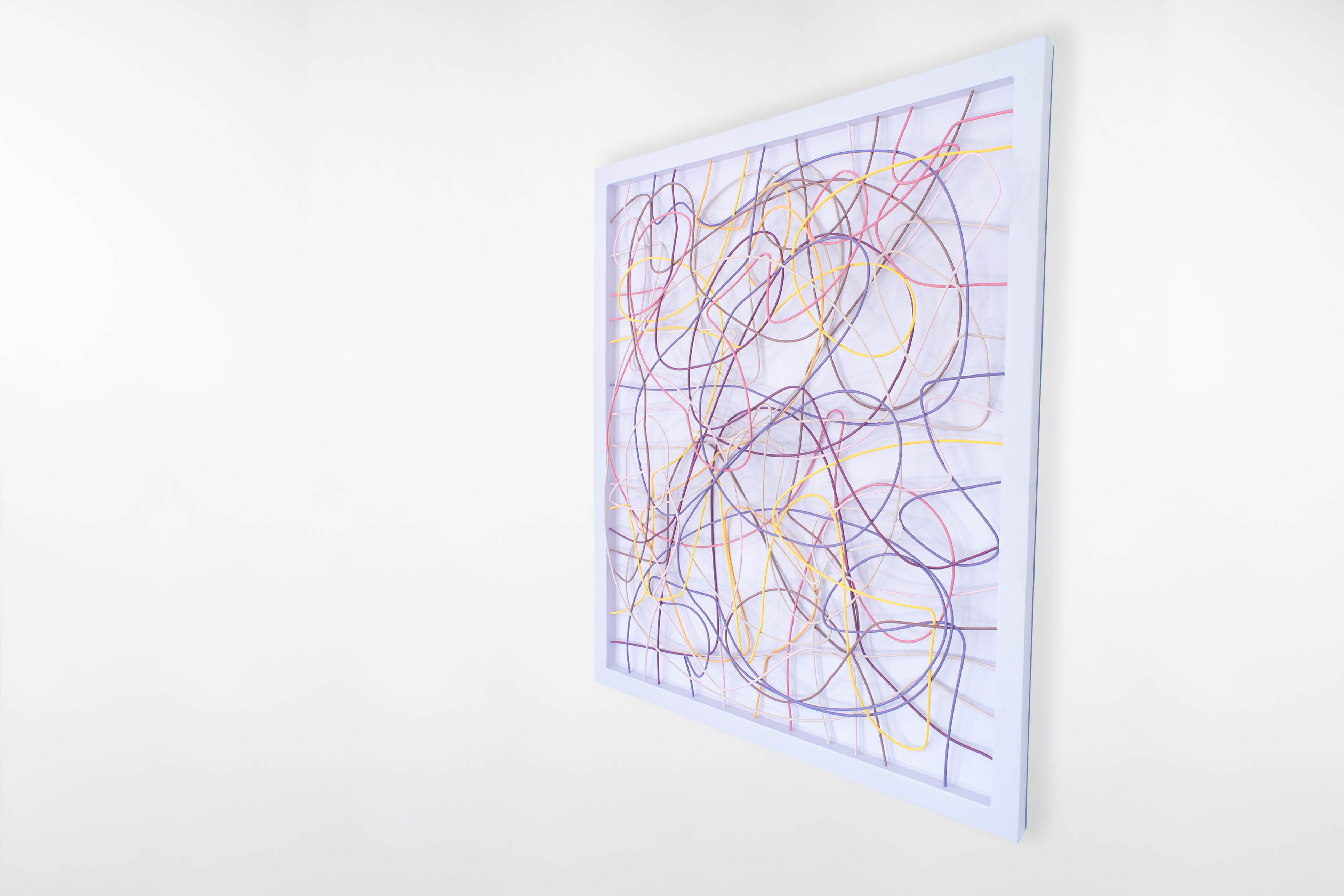 Side angle view of a Cameron Hunt Toyco series artwork featuring looping pastel wires in pink, yellow, purple, peach and brown inside a white frame on a white wall.