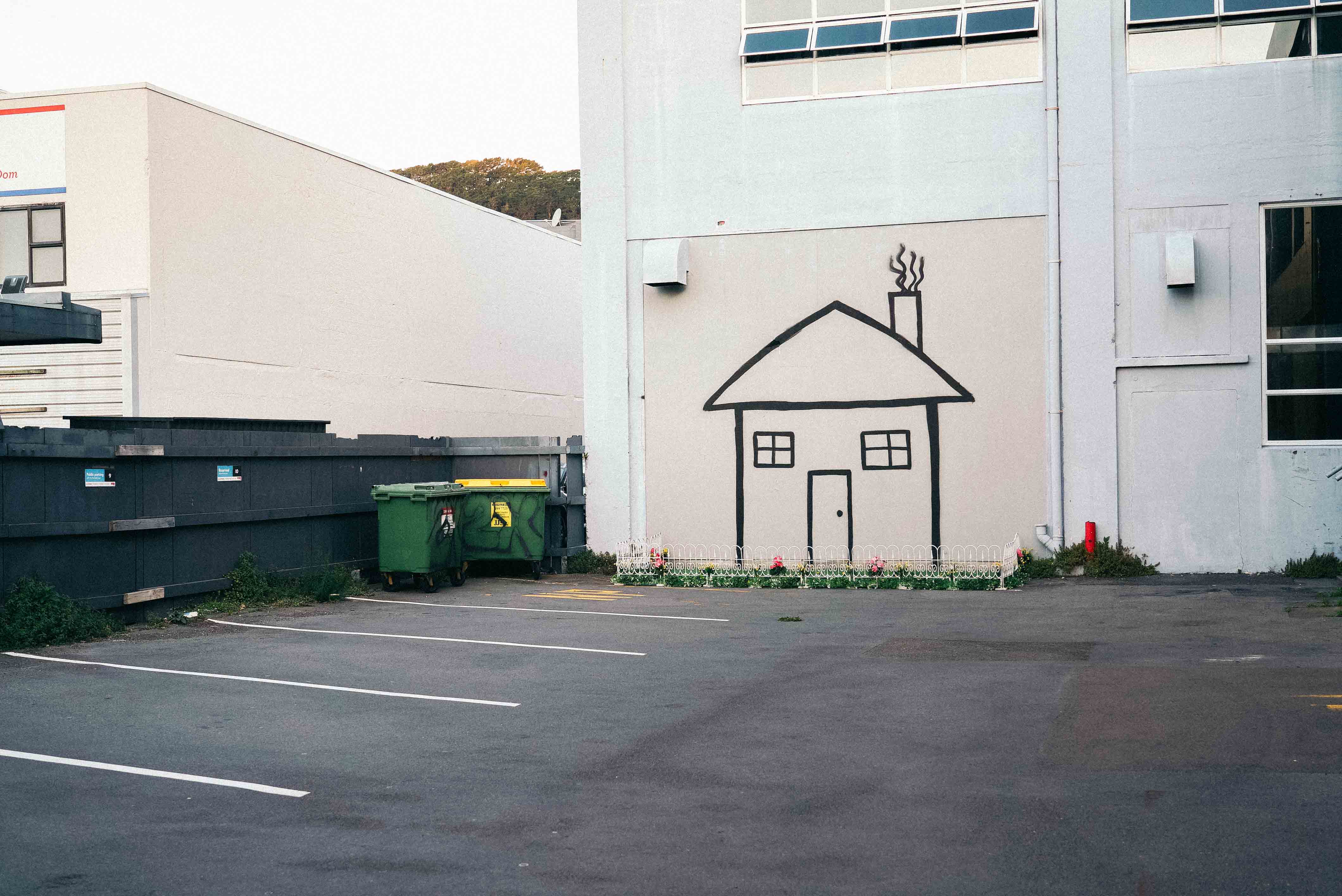 Street installation by Cameron Hunt featuring a simple black spray-painted outline of a cartoon-style house on the side of a building, complete with a white picket fence and artificial flower garden arranged along the base, playfully transforming a stark urban carpark.