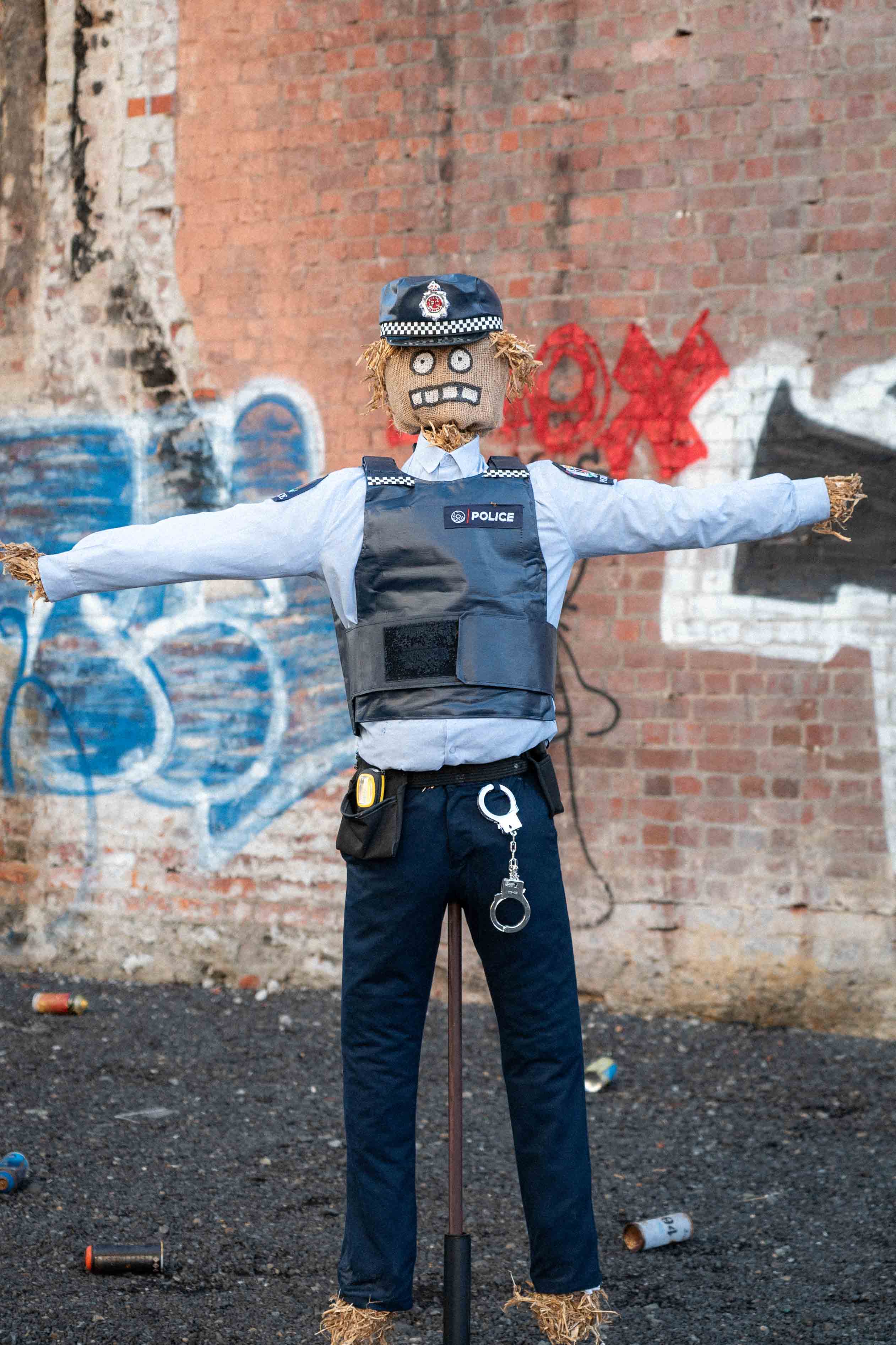 Street art installation by Cameron Hunt featuring a life-sized scarecrow dressed in a New Zealand police uniform, complete with high-vis vest, radio, handcuffs, and straw stuffing. Positioned against a graffitied brick wall, the scarecrow stands stiffly with outstretched arms, evoking a comical yet unsettling presence in an urban alleyway.