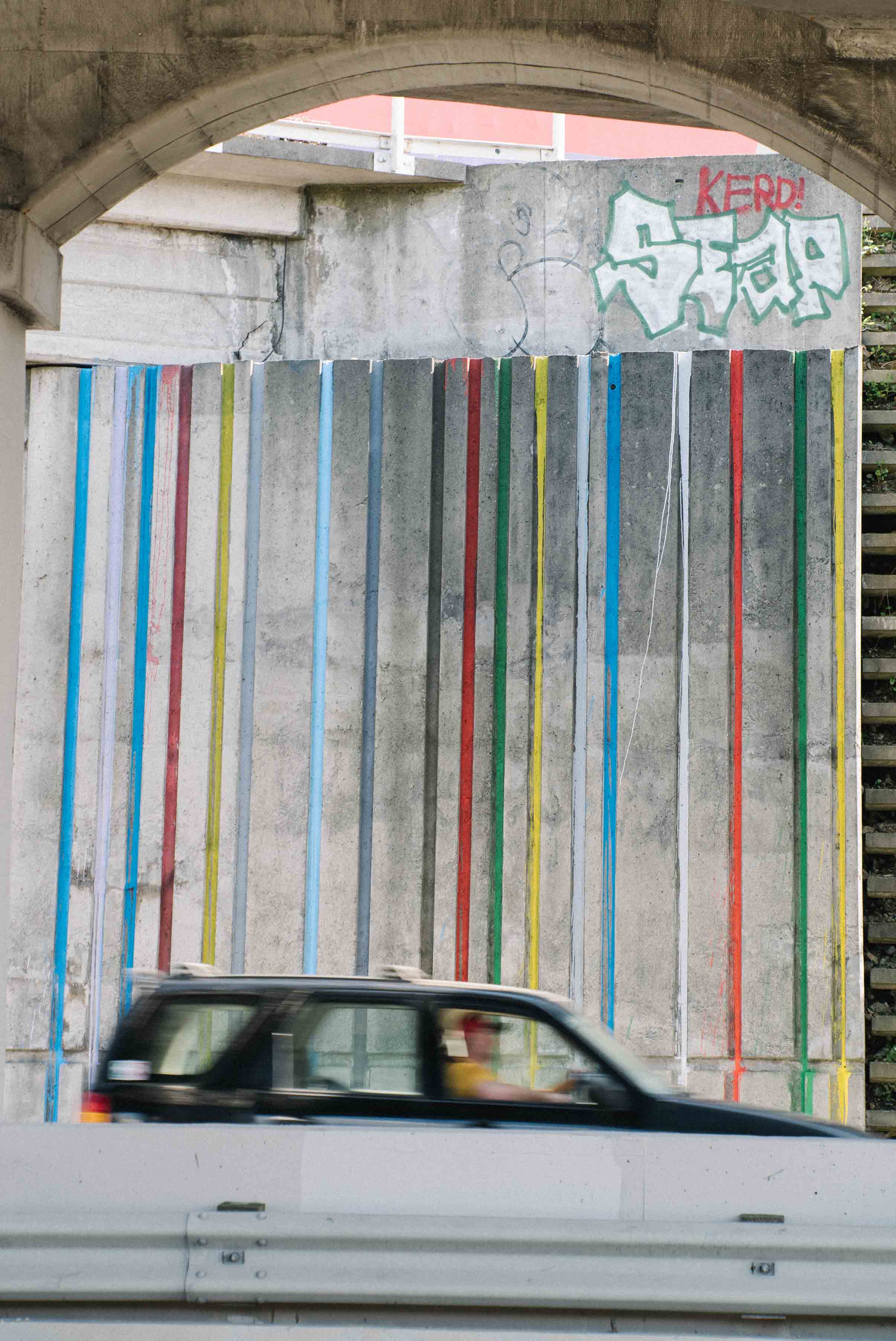 Street art installation by Cameron Hunt featuring vibrant drips of coloured paint—blue, red, green, yellow, and orange—poured down the vertical chutes of a concrete wall beneath an overpass, transforming a grey urban structure into a playful visual rhythm.
