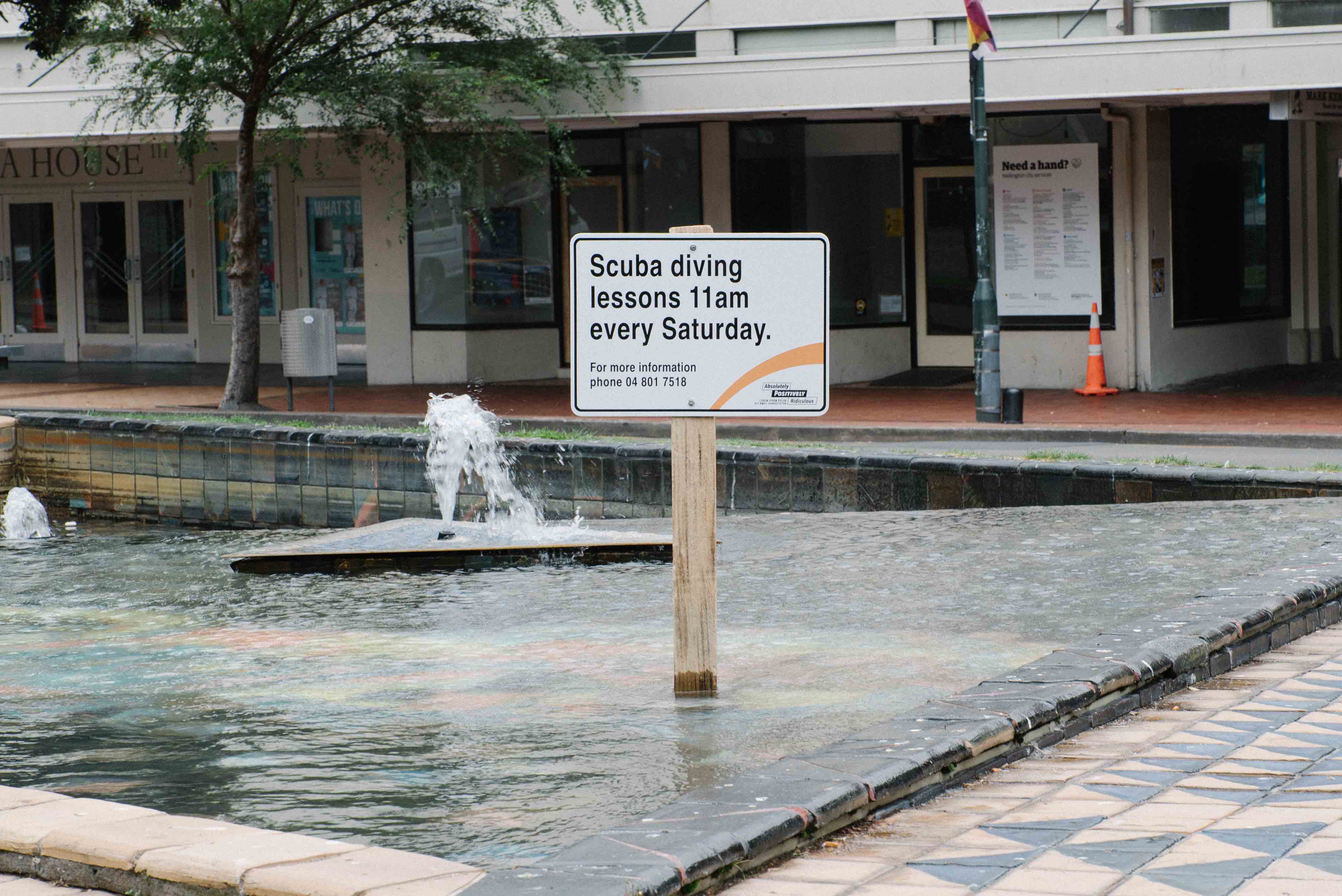Fake council sign installed in a public water fountain in Wellington, reading "Scuba diving lessons 11am every Saturday." Part of a street art intervention by Cameron Hunt, tricking passersby and officials.