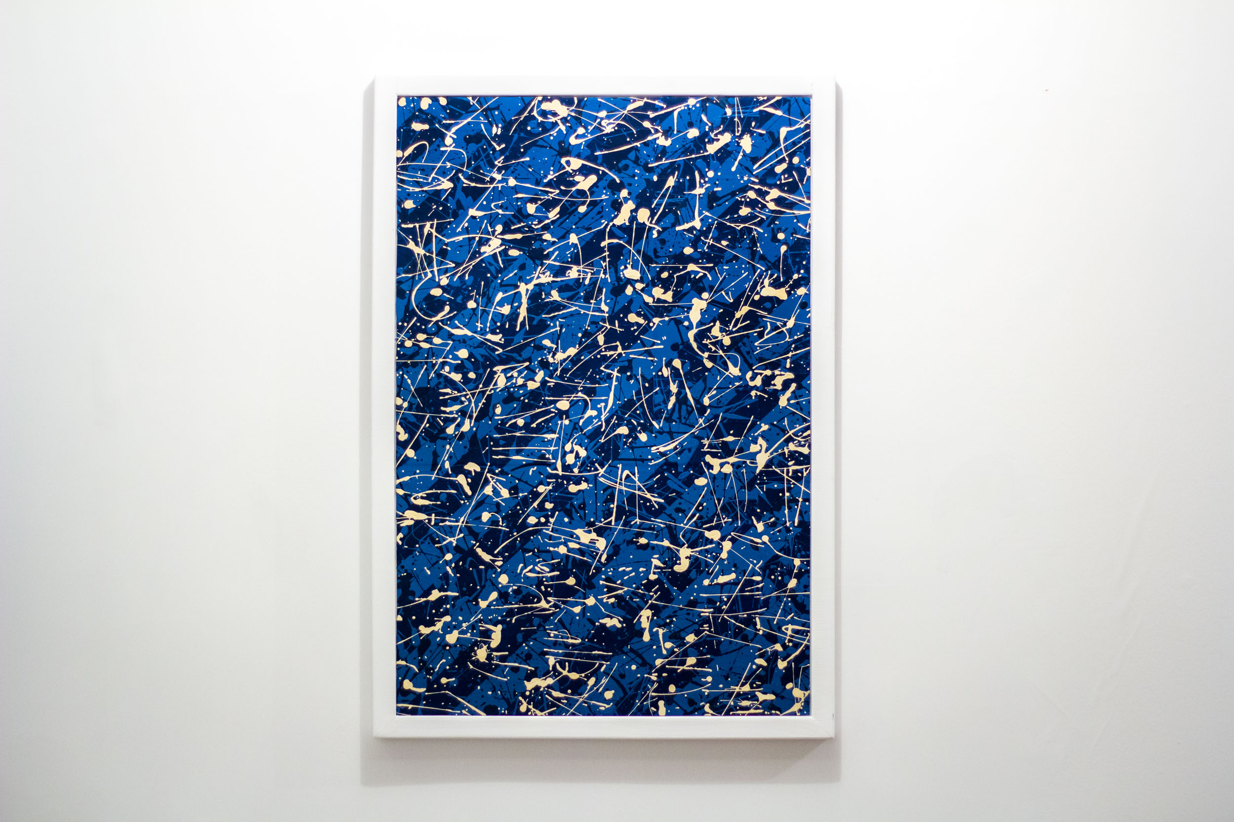 Framed abstract painting featuring layers of energetic cream-coloured splatters and sharp linear strokes over a deep blue background. The composition is dense and rhythmic, with a sense of movement and chaos. Set in a clean white rectangular frame.