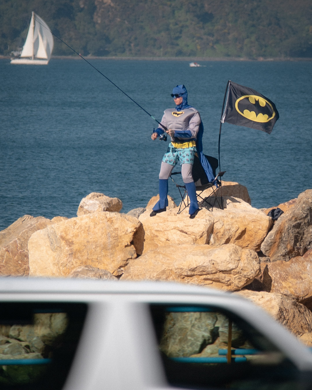 Street performance by Cameron Hunt dressed as Batman, complete with cape, utility belt, and swimming trunks, standing on coastal rocks beside a motorway. Holding a fishing rod and has planted a Batman flag beside him, while cars pass by in the foreground and sailboats drift in the background.
