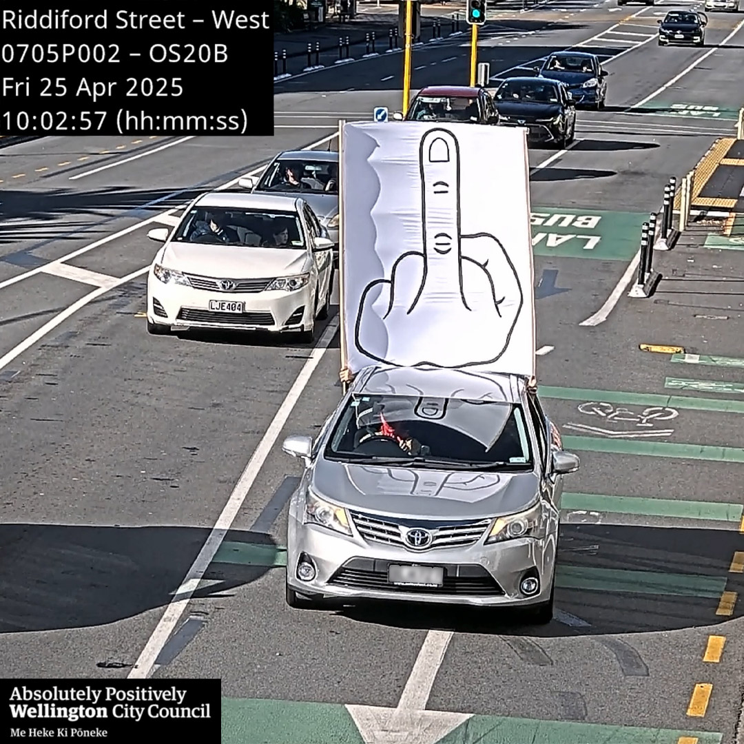 Street intervention by Cameron Hunt featuring a car driving through a bus lane with a large banner displaying a hand-drawn middle finger, intentionally triggering a traffic infringement camera to receive and repurpose the official council image as part of the artwork.