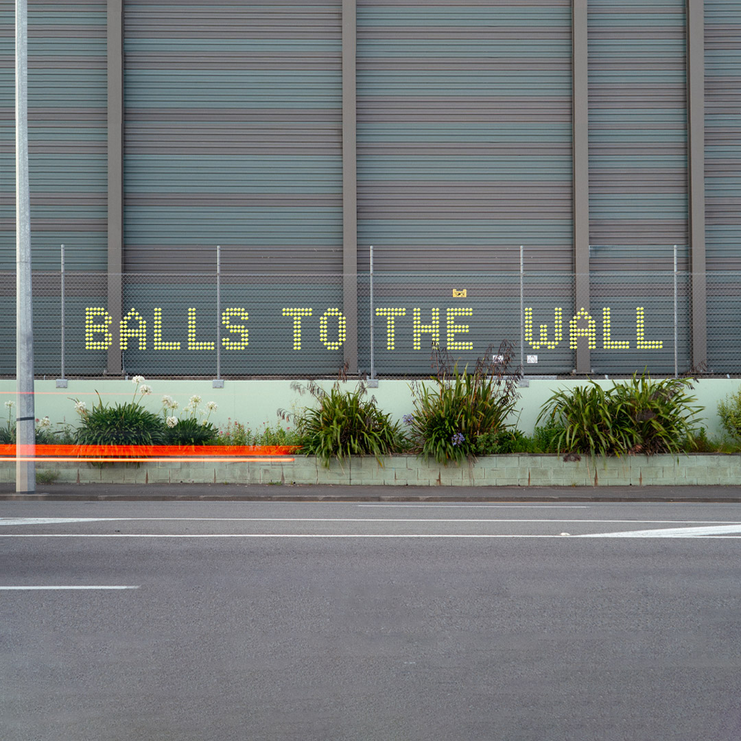 Street installation by Cameron Hunt, created using yellow tennis balls inserted into a wire fence alongside a busy urban street, forming bold text in a playful and provocative public artwork.
