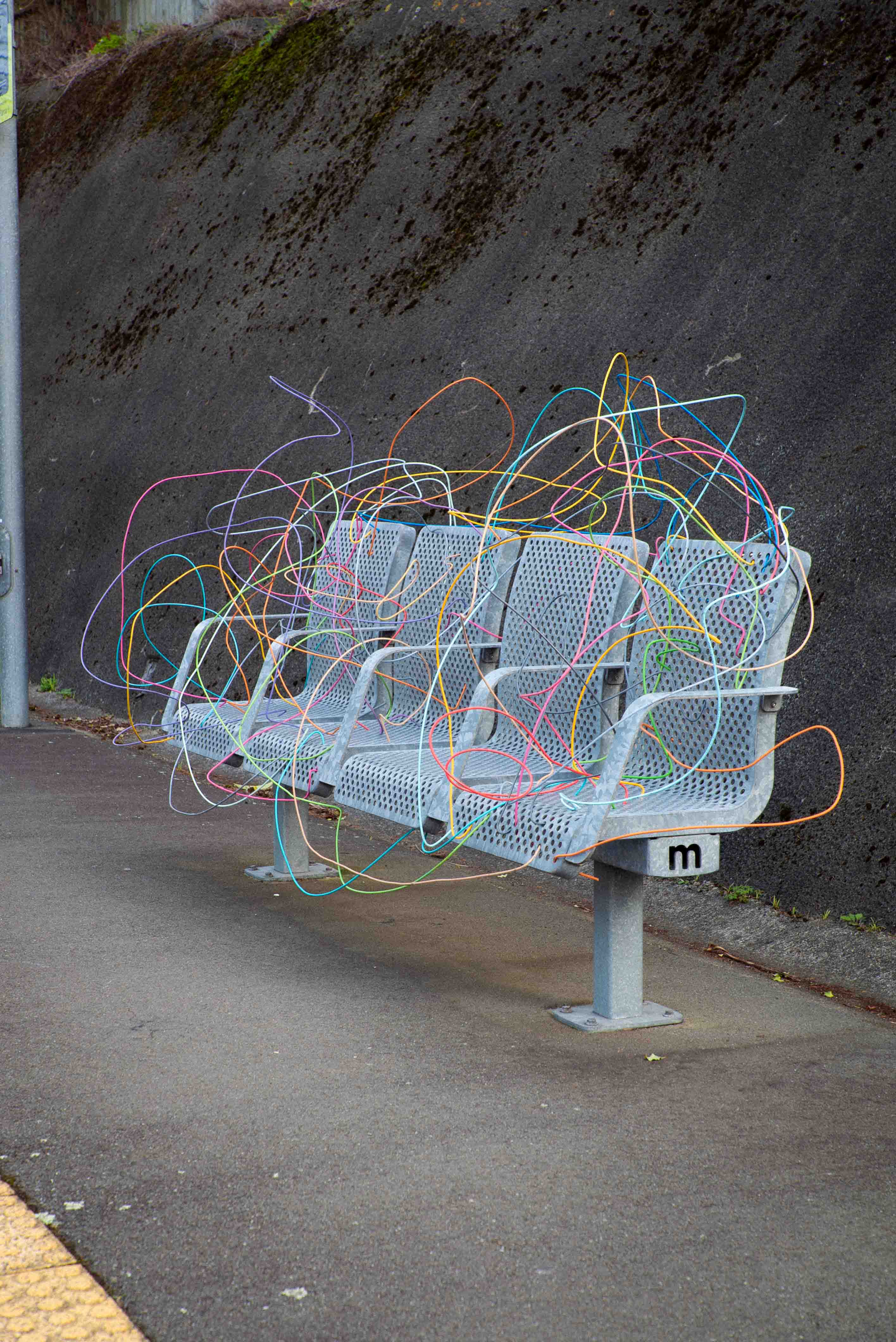 Street art installation by Cameron Hunt featuring a metal train station bench woven through with a chaotic explosion of multicoloured wires and plastic tubing. The vibrant, tangled mess disrupts the function of the bench while injecting playful energy into the drab grey setting of the station platform, contrasting order and disorder in public space.