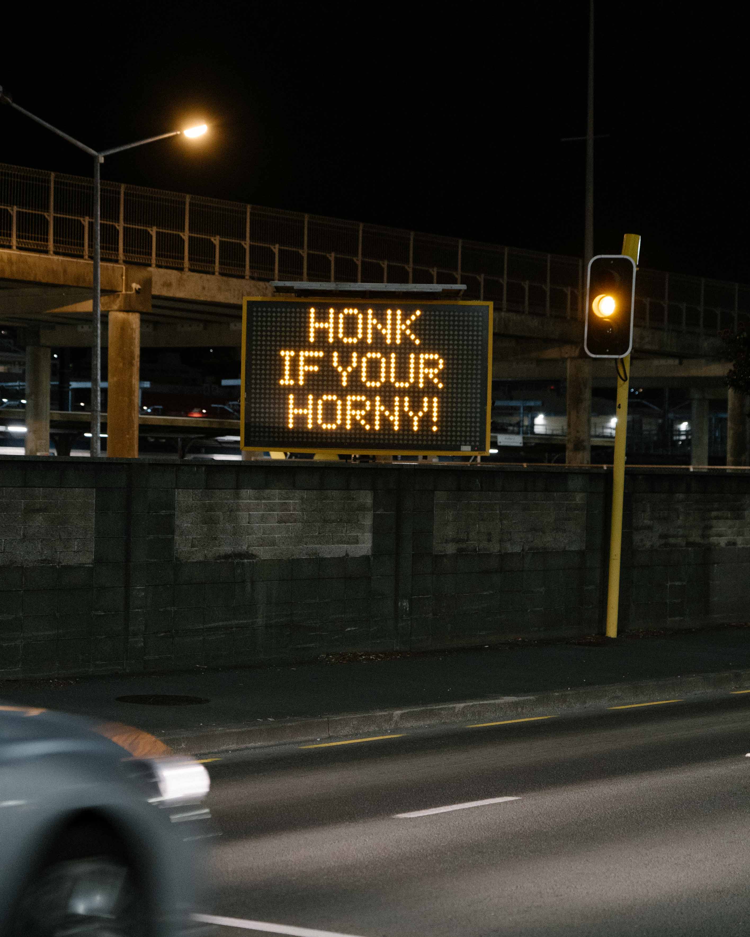 Humorous night-time street intervention by artist Cameron Hunt featuring a hacked electronic road sign reading "HONK IF YOU'RE HORNY!" on a Wellington roadside.