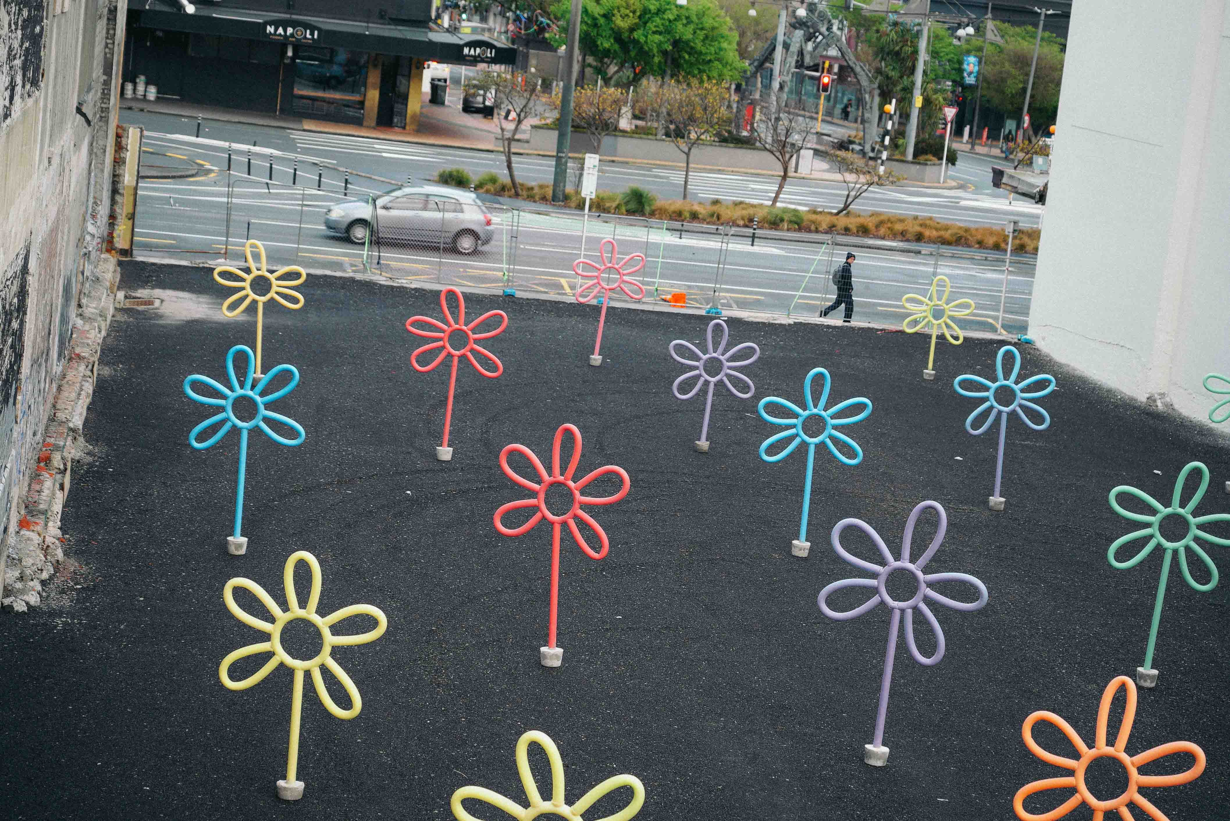 Street art installation by Cameron Hunt featuring a whimsical field of oversized flowers made from bent and fastened pool noodles in pastel shades of red, yellow, blue, green, orange, and purple, planted upright in an empty asphalt lot overlooking a city intersection.