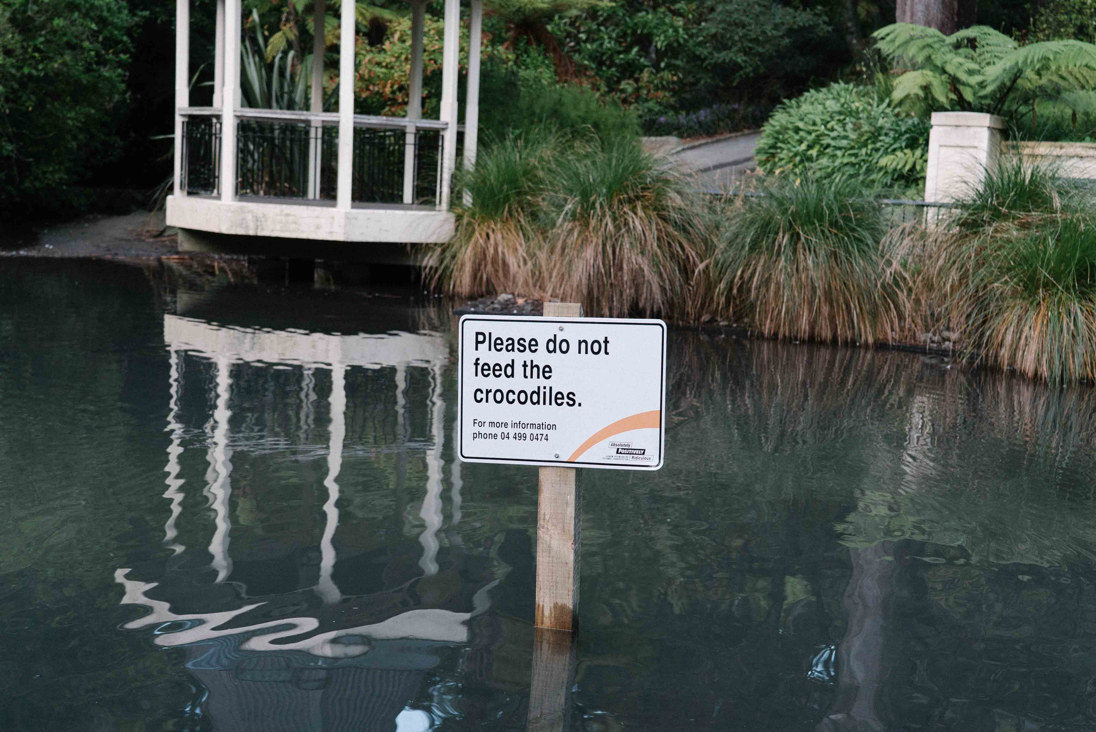 Playful fake council sign placed in a city pond reading "Please do not feed the crocodiles." Street art installation by Cameron Hunt designed to provoke surprise and amusement.