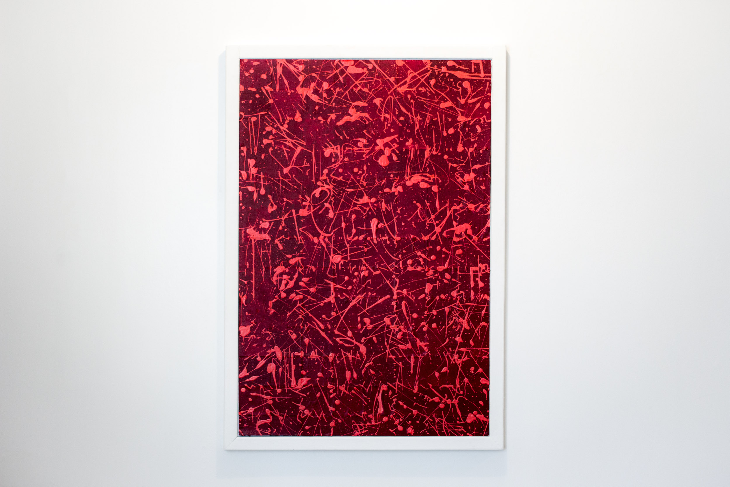 Framed abstract painting featuring vibrant red and pink paint splatters and fine linear strokes across a deep crimson background. The composition is dense and layered, creating a dynamic sense of movement and texture. Encased in a clean white rectangular frame.