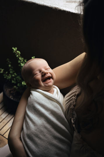 Smiling newborn baby wrapped in a white blanket and cradled in mother's arms during an in-home newborn photography session.