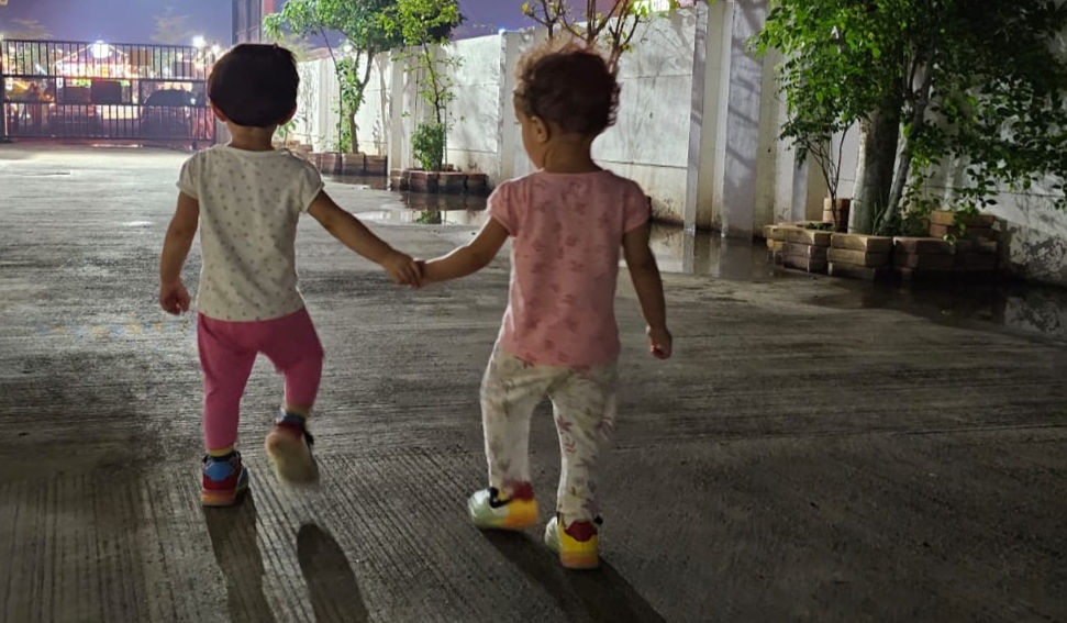 Two toddlers, one in pink pants and the other in a pink shirt, walk hand in hand on a dimly lit pathway lined with trees at night.