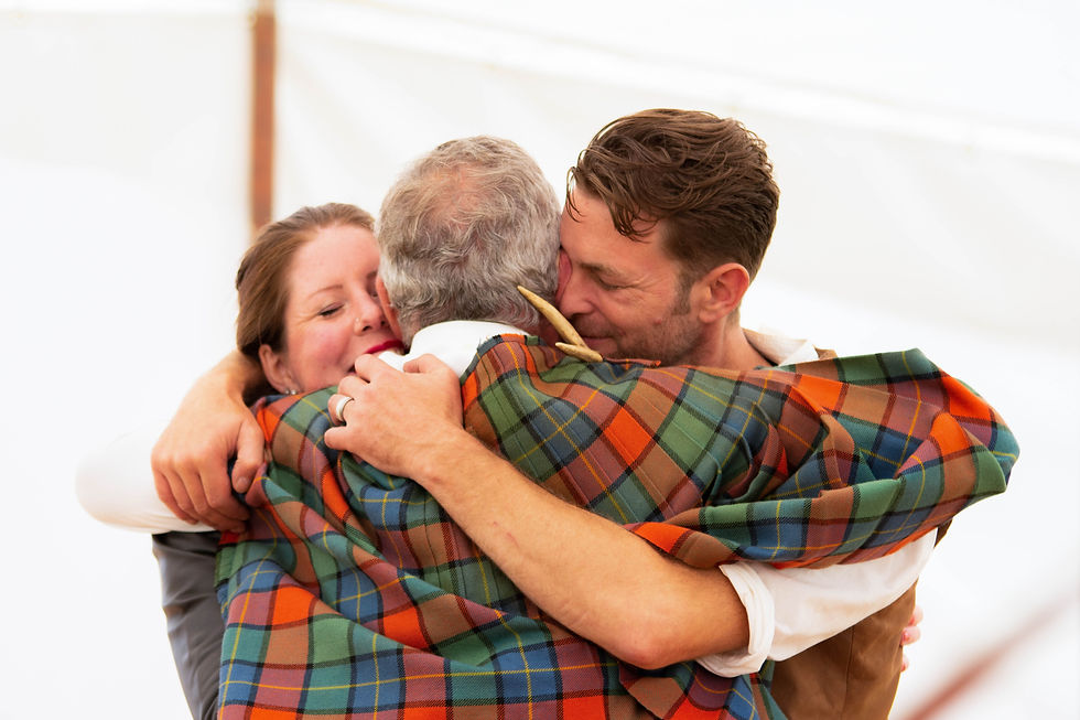 Three people share a warm embrace, draped in a colourful tartan cloak. The setting is bright and intimate, conveying a joyful mood.