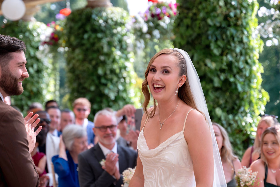 Bride in white dress smiling joyfully, standing with a groom in a suit. Guests clap in a garden setting with lush greenery and flowers.