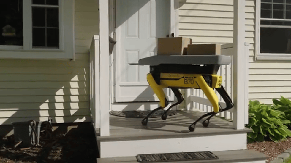 Yellow four-legged delivery robot labeled B70 carries boxes up a house porch in bright sunlight.