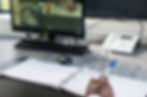 Close-up of a desk, mans hand is visible, writing in a notebook whilst looking at security monitor