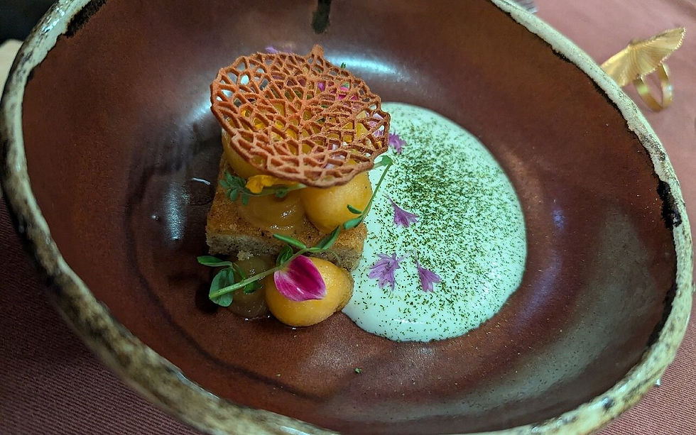 Gourmet dessert in a brown bowl; features a lattice garnish, orange dollops, sauce, and edible flowers on white cream with green herbs.