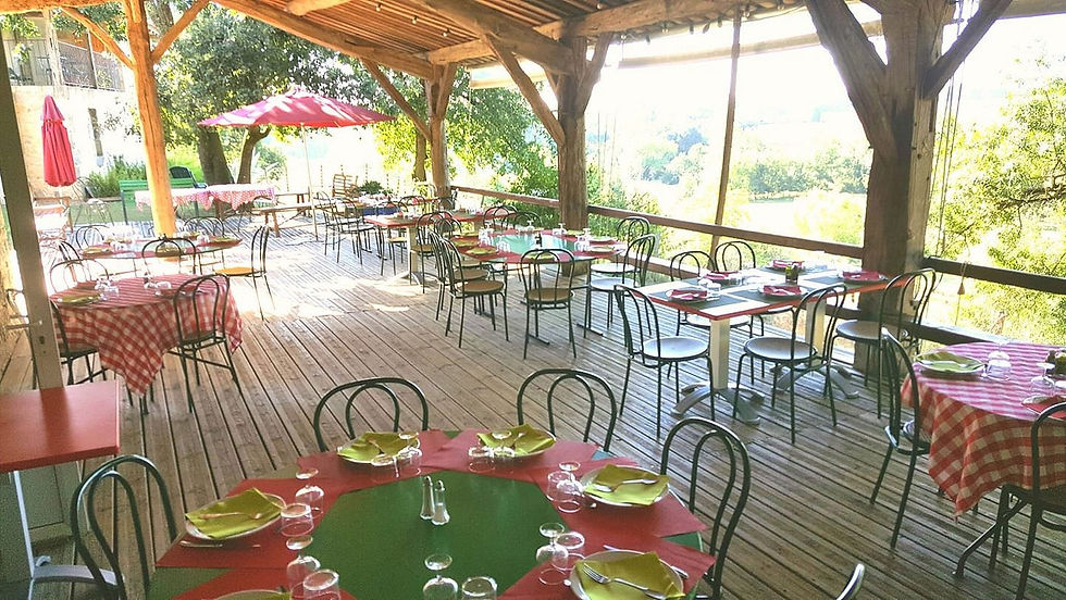 Outdoor patio with empty tables set with red and green tablecloths, yellow plates, and glasses. Red umbrella and trees in the background.