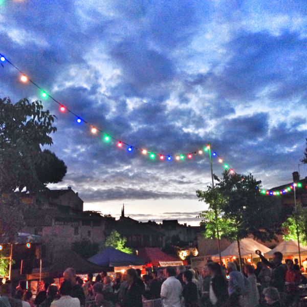 Crowd at an outdoor evening festival with colorful string lights under a cloudy sky. Tents and a lively atmosphere create a festive mood.