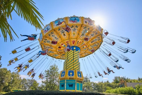 Colorful swing carousel spins with people enjoying the ride. Sun shines brightly, blue sky, green foliage. Joyful and lively atmosphere.