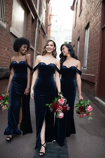 Three bridesmaids in dark velvet dresses with bouquets in an alley.