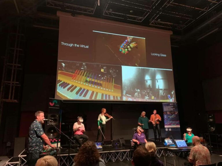 Musicians perform on stage with instruments; a large screen shows virtual instruments and the text "Through the Virtual" and "Looking Glass."