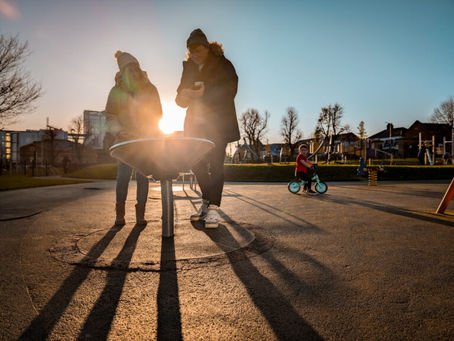 Two people stand at a playground table, backlit by the sun. A child rides a bike nearby. The setting is warm and inviting.