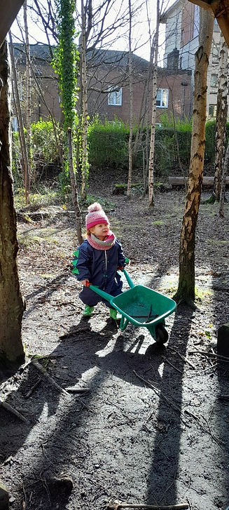 Child pushing a green toy wheelbarrow in a wooded garden.