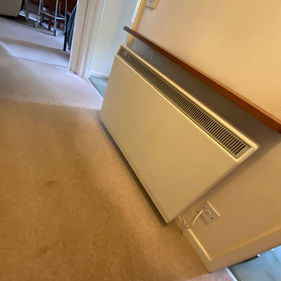 A white electric radiator mounted on a wall in a carpeted hallway, with a wooden shelf above it and a visible electrical outlet.